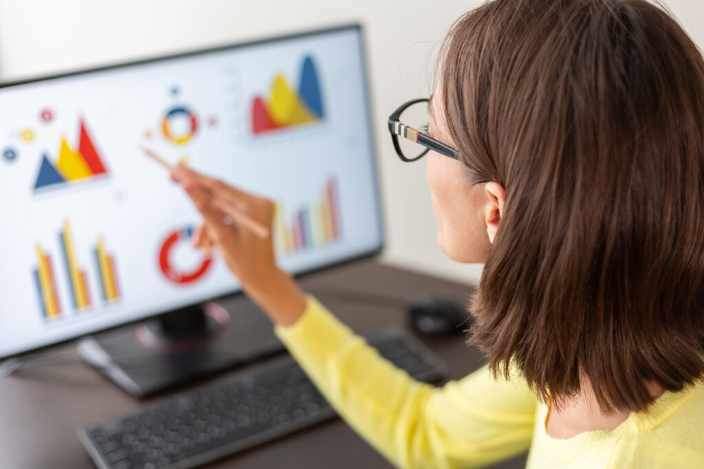 Kobieta w okularach analizuje grafy i wykresy na monitorze komputera, wskazując na dane palcem.