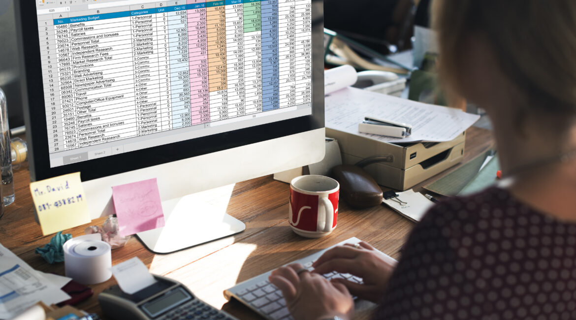 Kobieta pracująca na komputerze z otwartym arkuszem kalkulacyjnym Excel na monitorze, w biurze z widocznymi akcesoriami biurowymi.