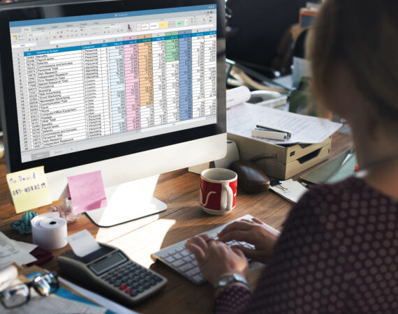 Kobieta pracująca na komputerze z otwartym arkuszem kalkulacyjnym Excel na monitorze, w biurze z widocznymi akcesoriami biurowymi.